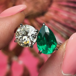 A hand with pink nails holds the Toi et Moi Pear Shape Emerald and Round Diamond Engagement Ring in White Gold, featuring a pear-shaped emerald and round diamond set in prongs, against a pink and brown blurred background.