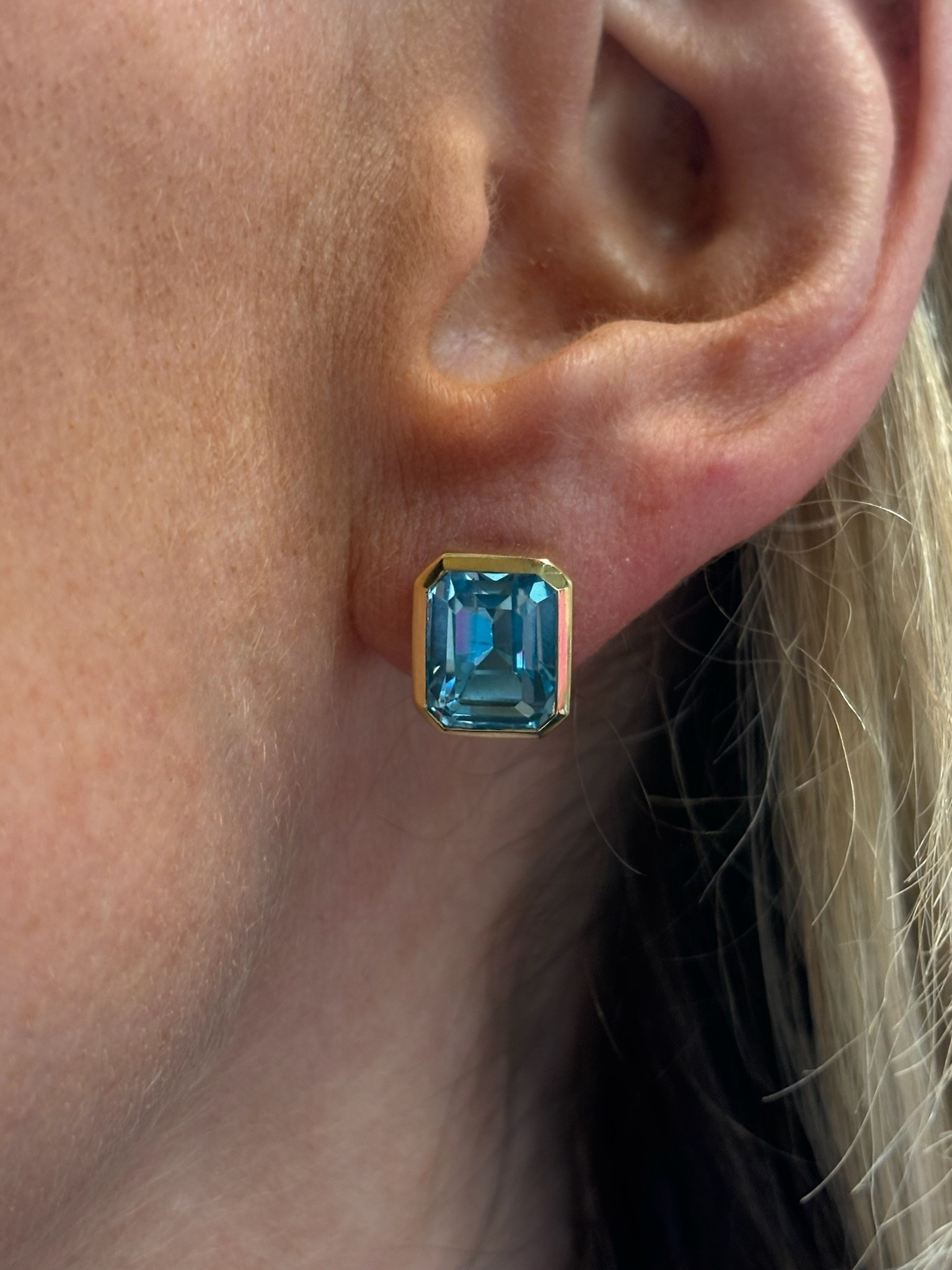 A close-up of an ear with light hair wearing the Bezel Set Emerald Cut Aquamarine Stud Earrings in Yellow Gold.