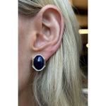 Close-up of a person with light blonde hair wearing Oval Lapis Lazuli Studs with Elongated Baguette Diamonds and Pavé—18K gold earrings featuring deep blue lapis lazuli surrounded by small baguette diamonds.