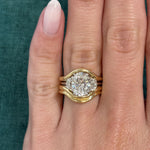 A manicured hand wears three stacked gold rings, including the Single and Double Band Contour Ring Jacket in Yellow Gold, with a central oval diamond flanked by two smaller diamonds, set against a green textured background.