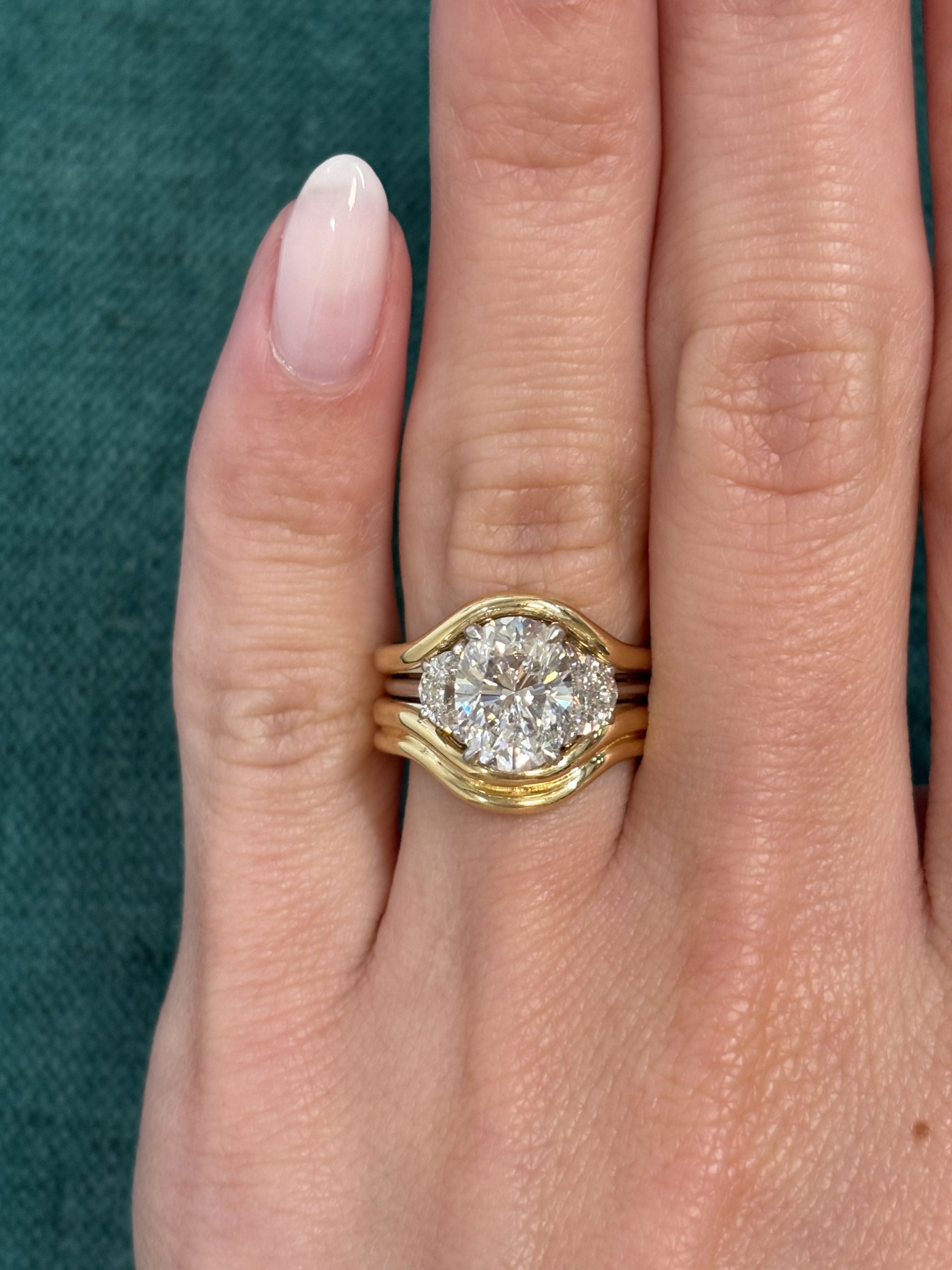 A manicured hand wears three stacked gold rings, including the Single and Double Band Contour Ring Jacket in Yellow Gold, with a central oval diamond flanked by two smaller diamonds, set against a green textured background.