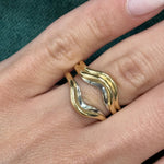 A close-up shows a hand wearing the Single and Double Band Contour Ring Jacket in Yellow Gold on the middle finger, with wavy yellow gold and silver bands fitting together like a puzzle against textured dark green fabric.