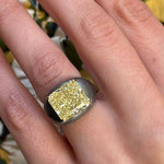 A hand wears the East-West Prong Set Radiant Cut Yellow Diamond Signet Ring in Black Rhodium, featuring a striking yellow diamond, against a floral-patterned background.
