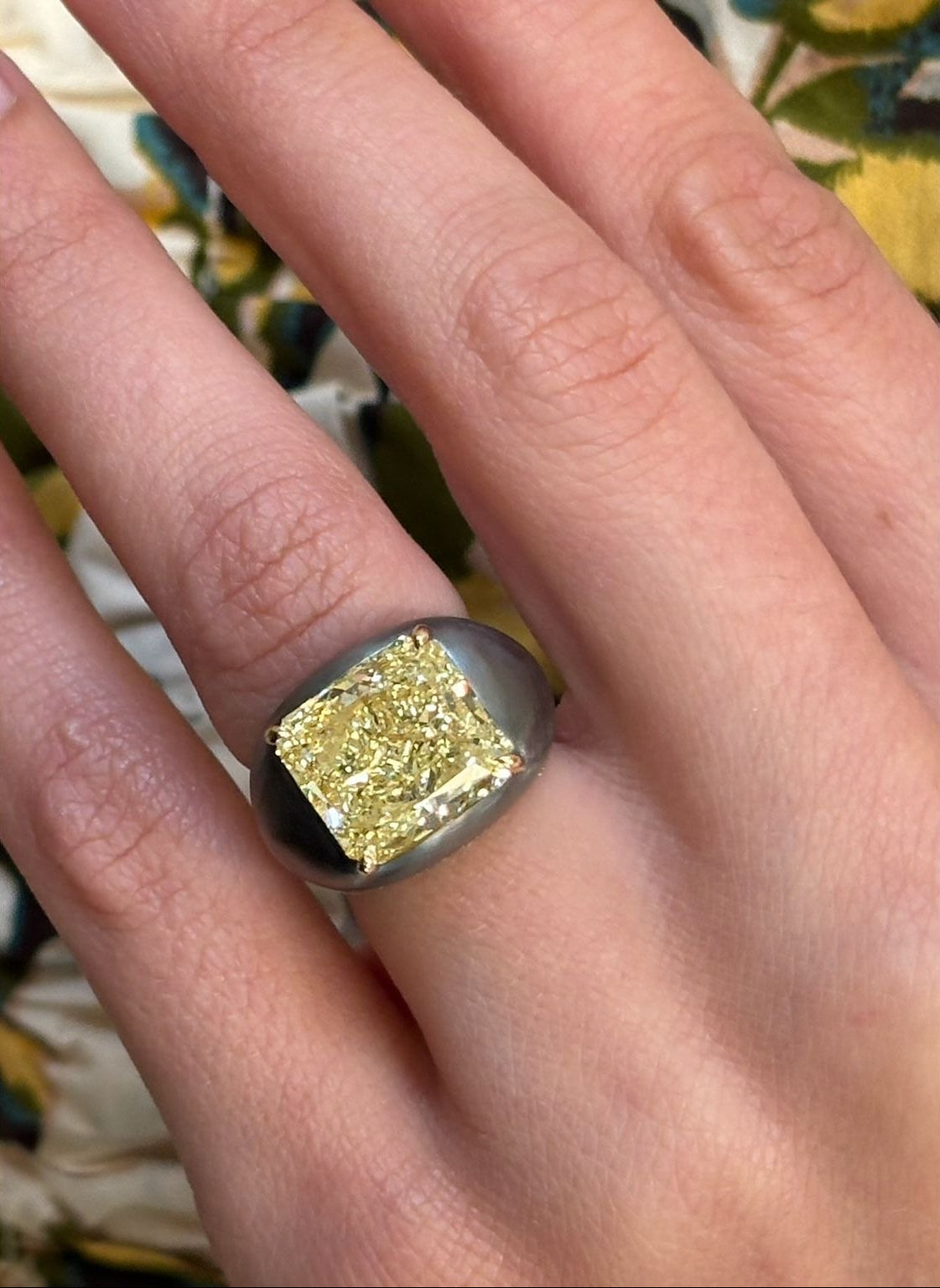 A hand wears the East-West Prong Set Radiant Cut Yellow Diamond Signet Ring in Black Rhodium, featuring a striking yellow diamond, against a floral-patterned background.