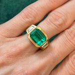 A hand wears the Bezel Set East-West Emerald Cut Emerald Three Stone Ring with Trapezoid Diamond Side Stones in Matte Yellow Gold, featuring a central emerald and two trapezoid diamonds, shown against a green background.