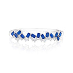 The Chroma Empire Alternating Emerald Cut Sapphire and Diamond Bangle in White Gold showcases alternating blue sapphires and diamonds in a sleek zigzag design, displayed on a reflective white surface.