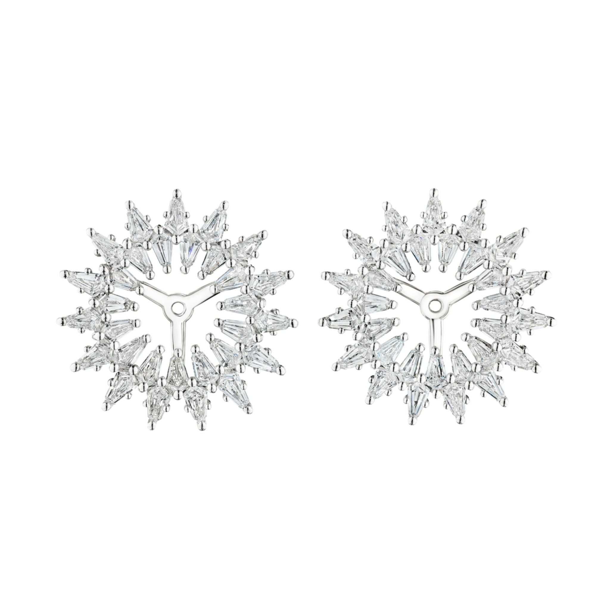 The Radial Shield Diamond Ear Jackets in White Gold are round, silver earrings with clear baguette-cut gemstones and shield cut diamonds arranged in a starburst pattern, resembling snowflakes.