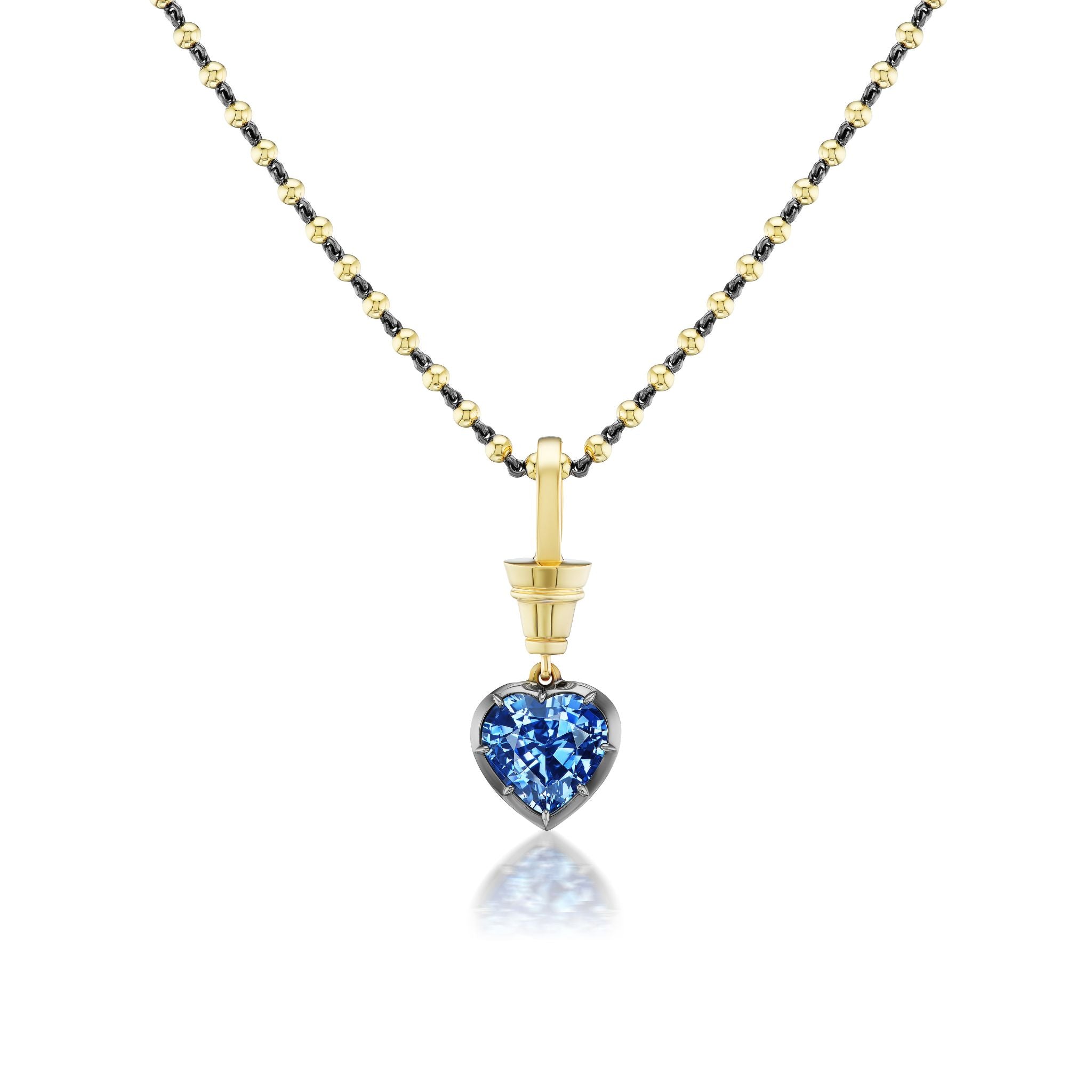 The Collet Set Heart Shape Blue Sapphire Necklace features a gold chain with black beads, a heart-shaped blue sapphire pendant in black rhodium and yellow gold, with its reflection shown on a white surface.