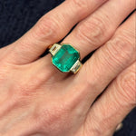 A hand showcases the Half Bezel Set Emerald Cut Emerald Engagement Ring with Trapezoid Diamond Side Stones in yellow gold, set against a dark, textured fabric background.