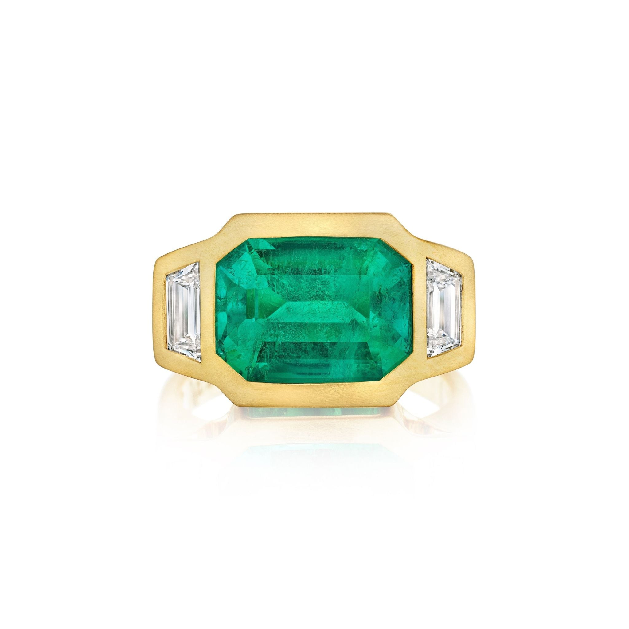 The Bezel Set East-West Emerald Cut Emerald Three Stone Ring features a center emerald and trapezoid diamond side stones, all set in matte yellow gold, shown against a white background for timeless elegance.