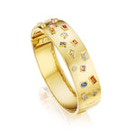 The Bezel Set Multi Shape Mixed Stone Bangle in Yellow Gold features diamonds, rubies, sapphires, and citrines arranged in geometric patterns across its polished surface.