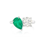 The Toi et Moi Pear Shape Emerald and Round Diamond Engagement Ring in White Gold features a striking silver band that displays a pear-shaped emerald alongside a round diamond, both elegantly set in prongs against a pristine white gold background.