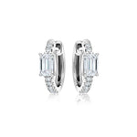 The Pavé Huggie with Emerald Cut Diamonds features silver hoops, each centered with an emerald cut diamond and accented by smaller round gemstones along the front.