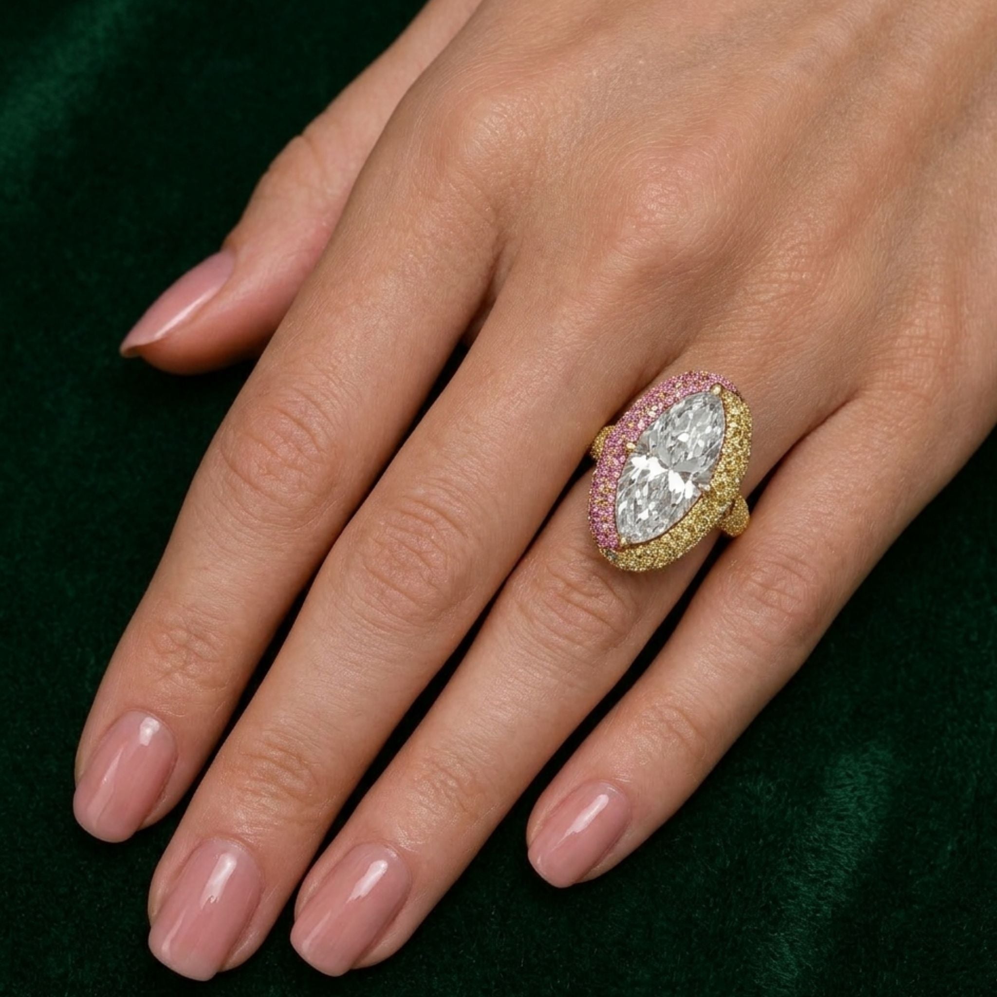 A hand with manicured nails showcases the Colorful Cobblestone Pavé Marquise Diamond Bombé Engagement Ring in yellow gold, featuring a marquise center and vibrant pink and yellow gemstones, against a dark green background.
