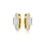 The Pavé Huggie with Emerald Cut Diamonds features 18k gold, small round pavé diamonds, and a larger rectangular emerald-cut diamond on each earring, shown on a white background.