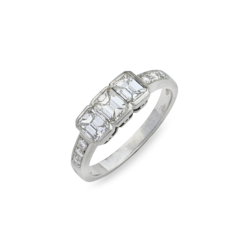 The Three-Stone Diamond Ring showcases three rectangular diamonds set side by side on a platinum band, accented with smaller stones along the sides. Displayed on a white background.