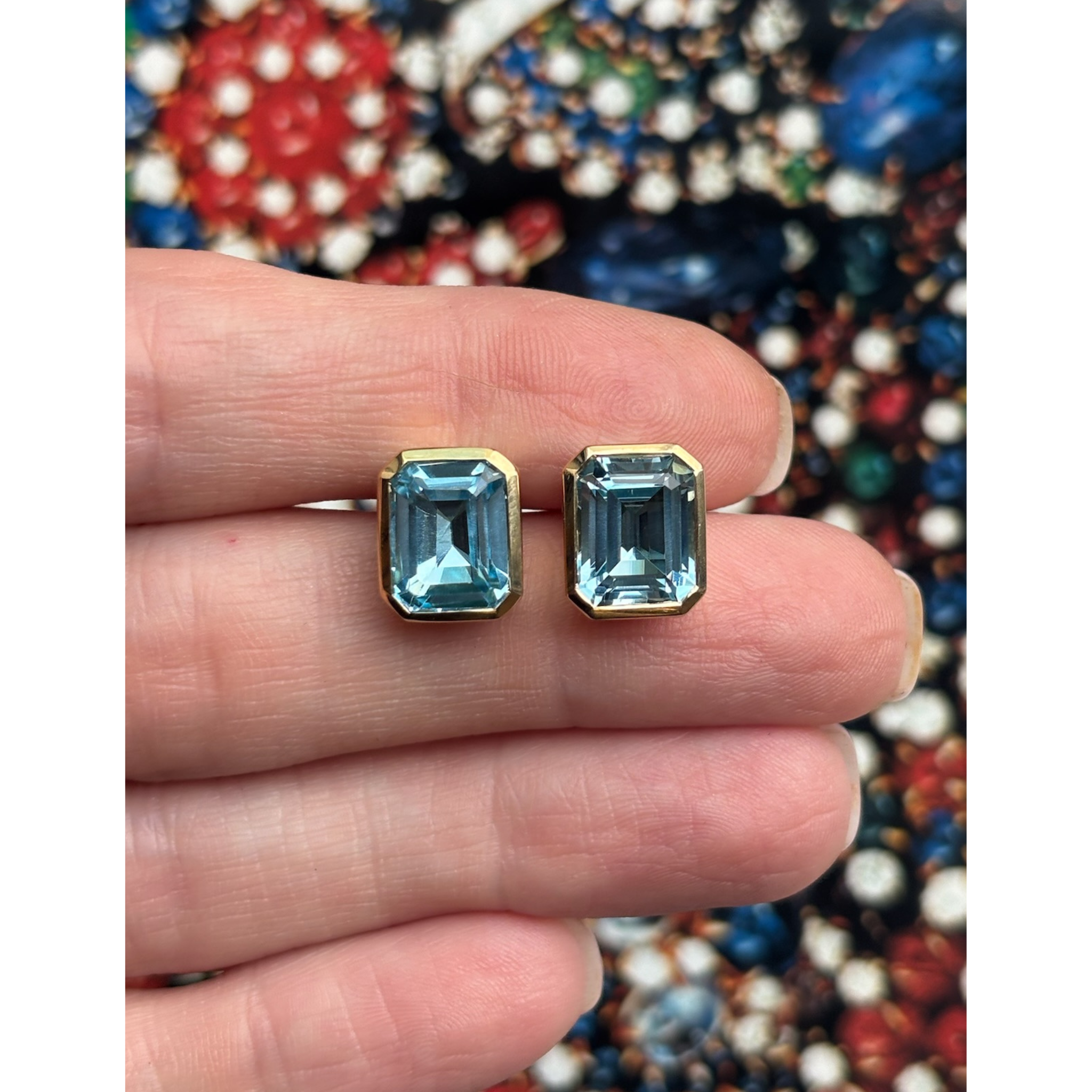 A hand displays the Bezel Set Emerald Cut Aquamarine Stud Earrings in Yellow Gold against a vibrant, jeweled backdrop.