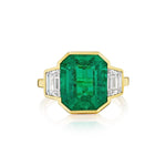 Introducing the Half Bezel Set Emerald Cut Emerald Engagement Ring with Trapezoid Diamond Side Stones in Yellow Gold—an exquisite ring crafted in 18k yellow gold, showcasing a striking emerald-cut green emerald center stone accented by two elegant trapezoid diamonds on each side.