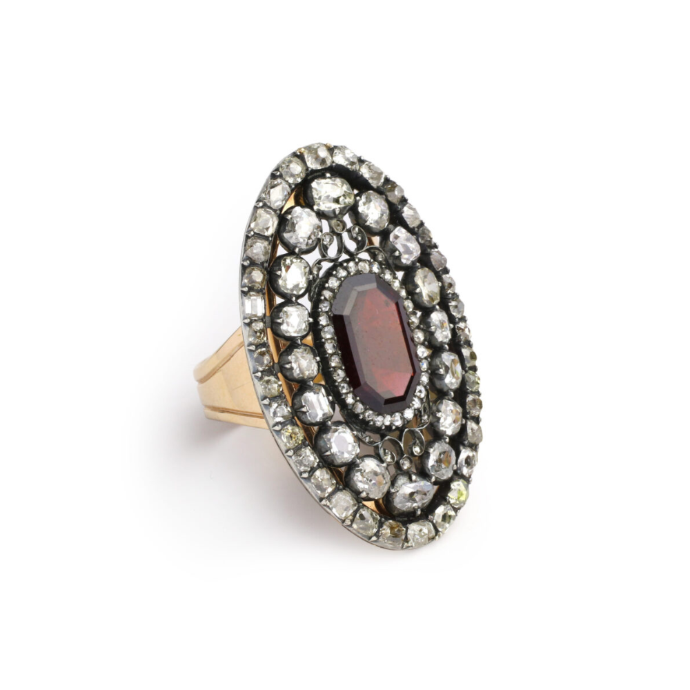 The Antique Garnet and Diamond Ring showcases an elongated oval design with a central cushion-cut garnet, accented by small old-cut diamonds in an ornate raised setting.