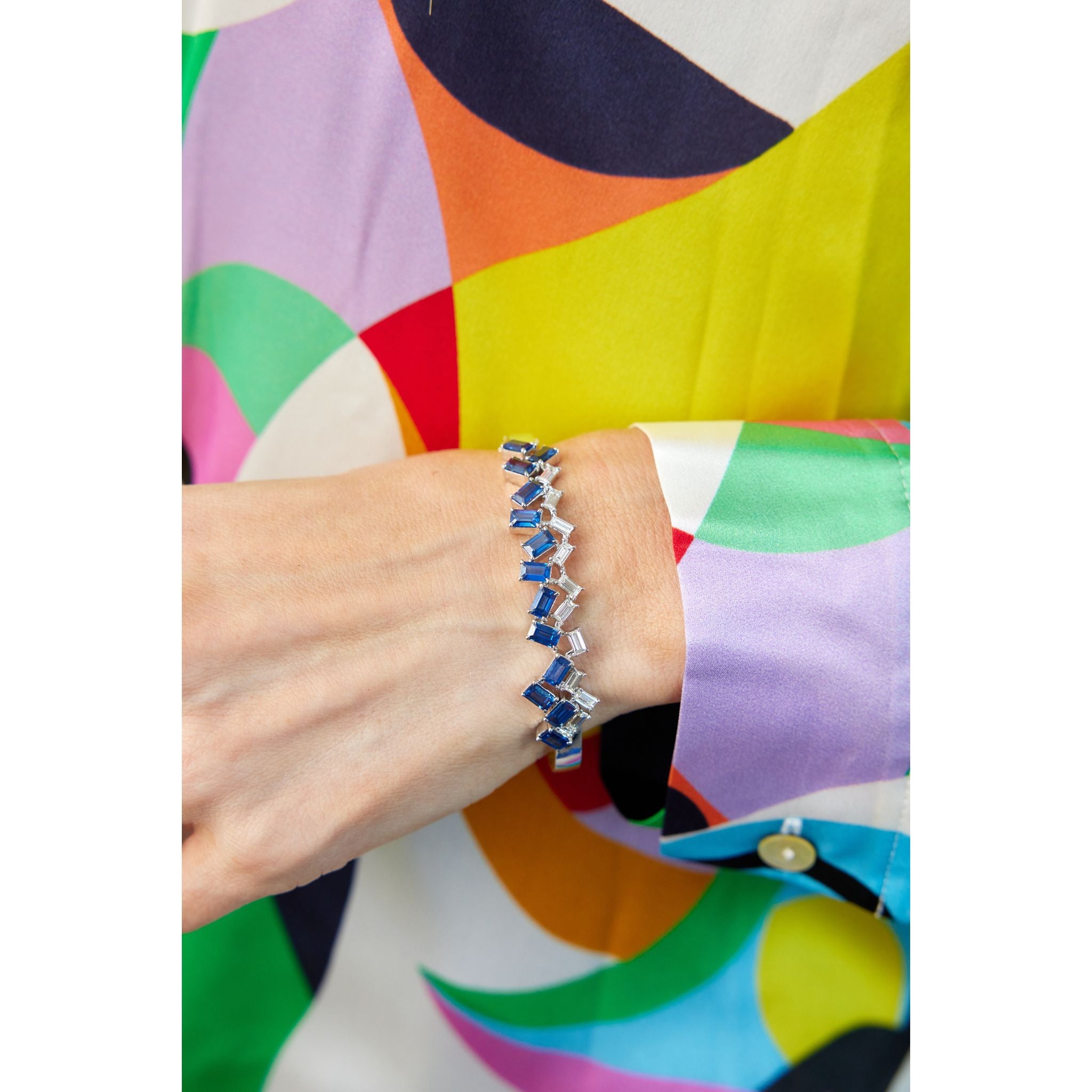A person in a colorful, geometric-patterned shirt showcases the Chroma Empire Alternating Emerald Cut Sapphire and Diamond Bangle in white gold, featuring blue sapphires and sparkling diamonds on their wrist.