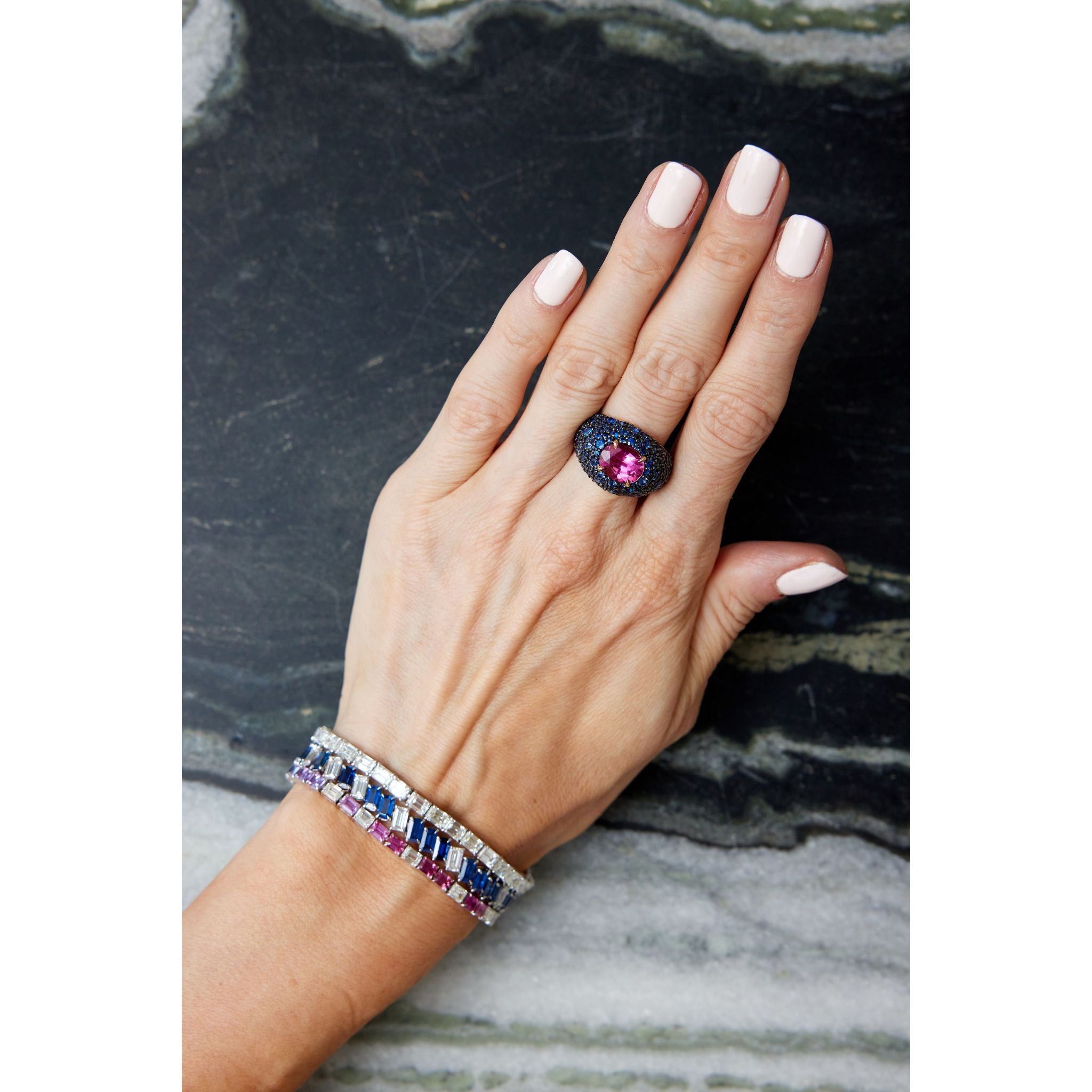 A hand with light pink nail polish rests on dark marble, adorned with a black ring featuring a large pink stone, two beaded bracelets with silver and multicolored gemstones, and the Chroma Empire Diagonally Set Emerald Cut Sapphire and Diamond Tennis Bracelet.