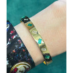 The Alternating Emerald Cut Diamond and Emerald Bangle in Yellow Gold is worn on a wrist with a dark, patterned long-sleeve shirt against a teal background.
