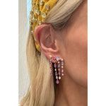 A close-up shows a woman's ear wearing Multicolor Waterfall Earrings with mixed shape sapphires and diamonds, set in 18K yellow gold. She also wears a yellow headband adorned with large, reflective gems. Only part of her face is visible.