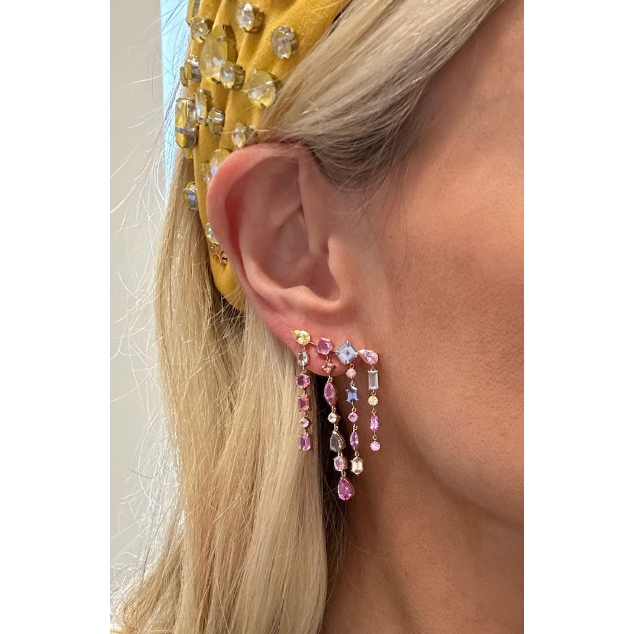 A close-up shows a woman's ear wearing Multicolor Waterfall Earrings with mixed shape sapphires and diamonds, set in 18K yellow gold. She also wears a yellow headband adorned with large, reflective gems. Only part of her face is visible.
