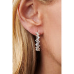 Close-up of a woman's ear wearing Zigzag Emerald Cut Diamond Hoops in 18K white gold, featuring rectangular and round diamonds in a zigzag pattern, with blonde hair partially visible.