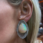 A close-up of a person’s ear adorned with a Vintage Opal and Diamond Earring, featuring a large teardrop-shaped opal encircled by platinum and diamond accents. The earring shimmers with iridescent colors, evoking the elegance of a diamond waterfall. The wearer has light skin and blonde hair.