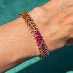 A wrist showcases the Half Bezel Set Rainbow Sapphire Chevron Tennis Bracelet in rose gold, featuring interlocking multicolor sapphires in a chevron design, set against a teal background.