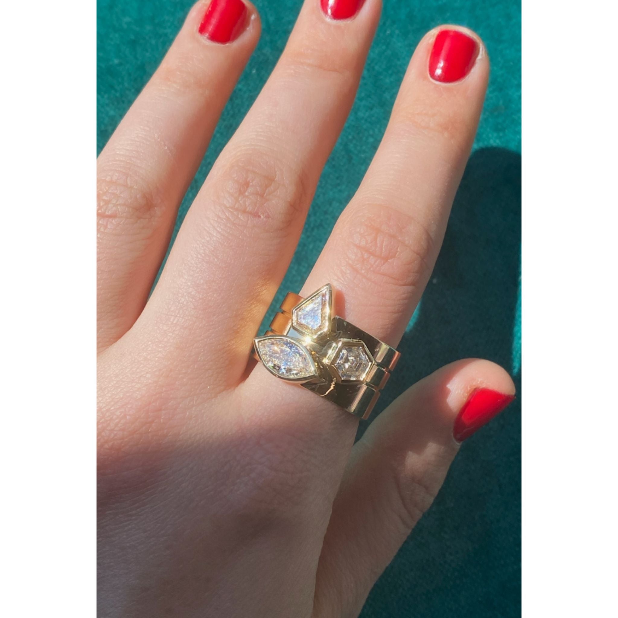 A hand with red nails wears the Bezel Set Mixed Shape Diamond Thick Band in Yellow Gold, featuring two geometric bezel-set diamonds that sparkle against a teal background.