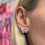 Close-up of a woman's ear wearing a Pair of Diamond Earclips featuring marquise diamonds and pear-shaped clear gemstones in a curved 18K white gold design. The background is softly blurred in pink and red tones.
