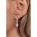 Close-up of a person's ear wearing Statement Emerald, Baguette, and Round Cut Diamond Drop Earrings in White Gold. The earrings have geometric shapes with three clear stones and baguette accents. Strands of light brown hair are visible.
