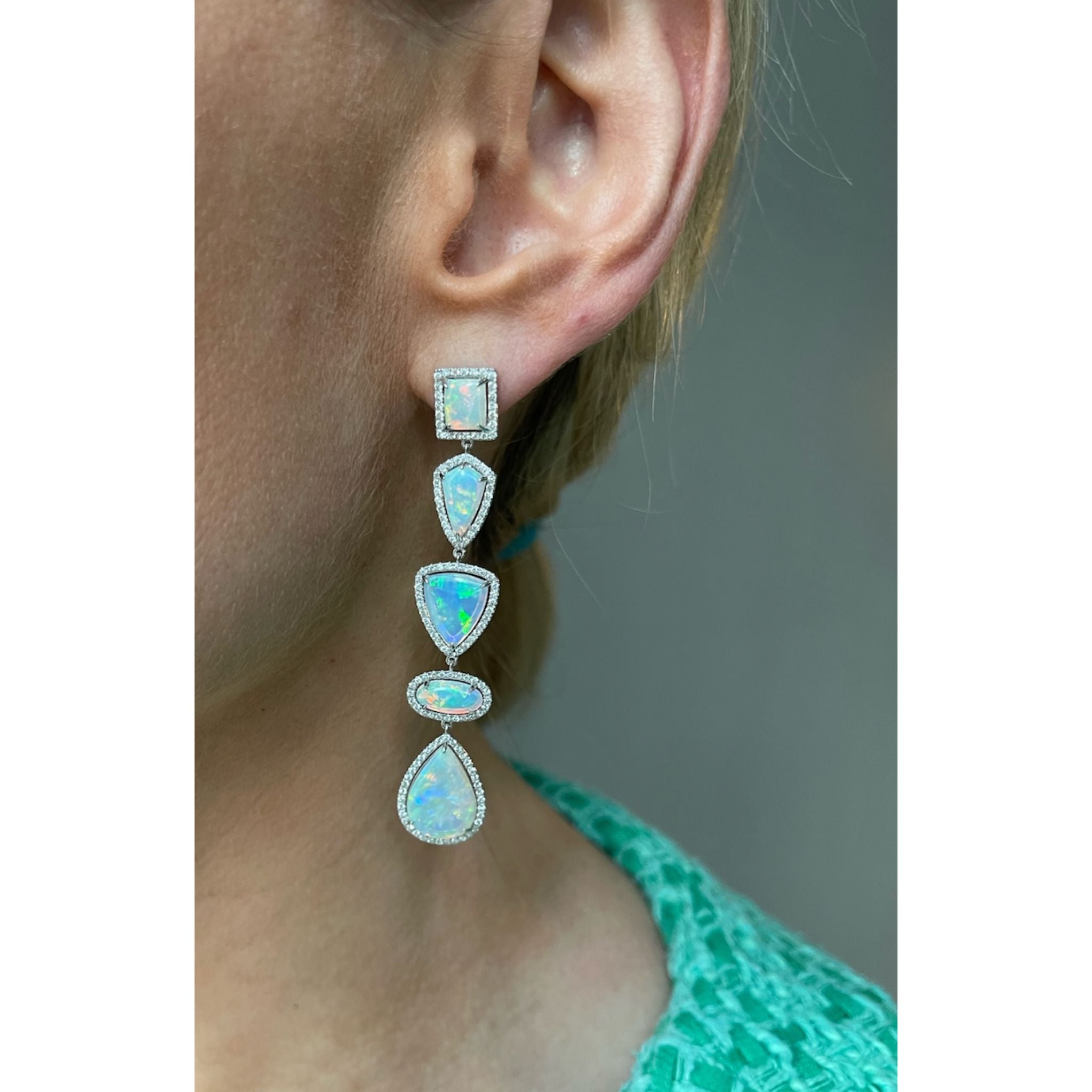 A close-up of a woman's ear wearing Opal Chandelier Earrings in 18K white gold, with four geometric opals outlined by clear stones. She is in a textured green top.