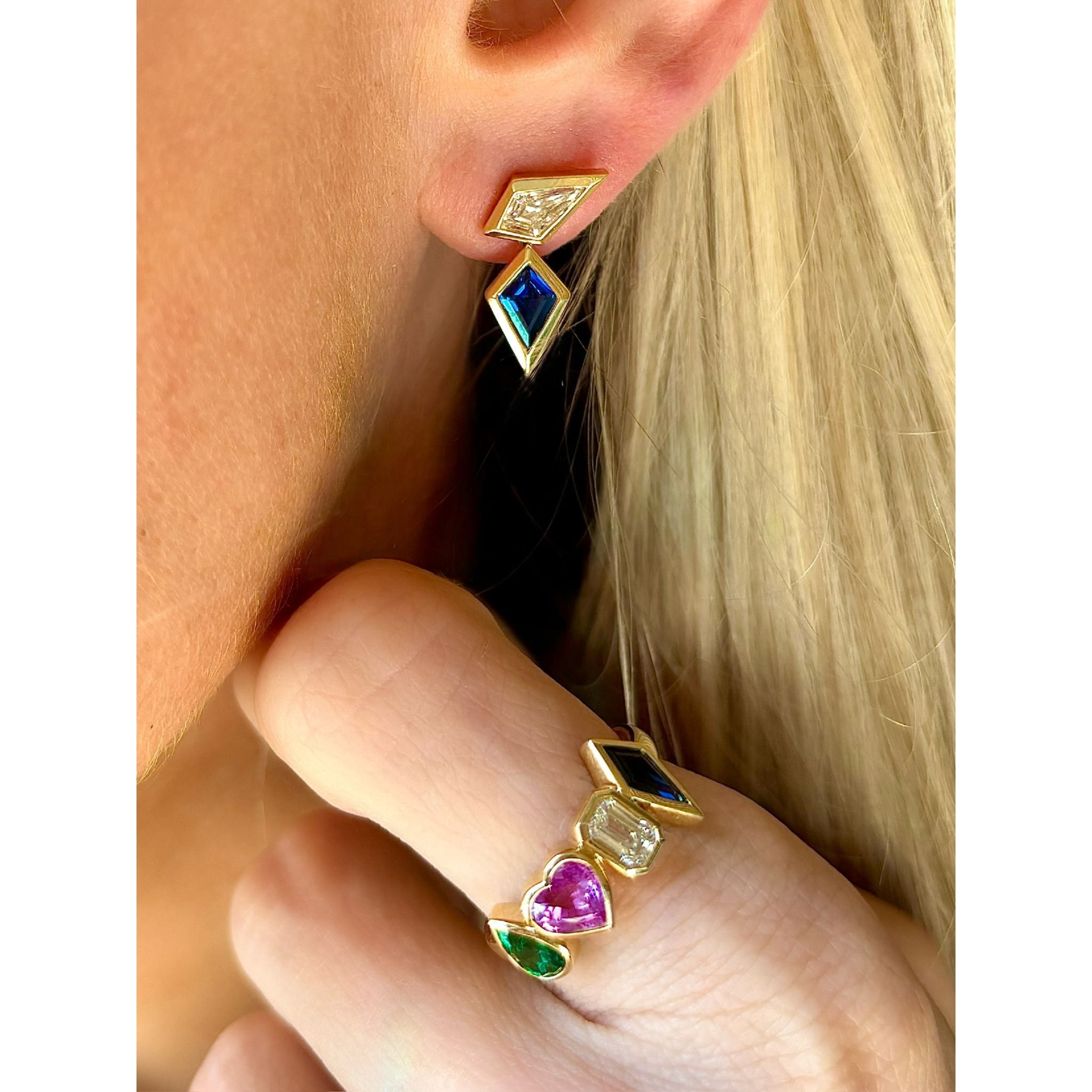 Close-up of a person wearing geometric gold earrings and the Chroma Heart Beats Bezel Set Cluster Ring in yellow gold, featuring mixed shaped emeralds, diamonds, and sapphires, as their hand gently touches their face.