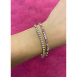 A wrist adorned with a Multicolor Asscher Cut Diamond and Sapphire Tennis Bracelet in Yellow Gold sparkles against a vivid pink textured background.