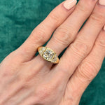 A hand with manicured nails wears the Burnish Set Cushion Diamond Engagement Ring with Half Moon Side Stones, showcasing a large cushion cut diamond in a bezel setting at the center, flanked by sparkling half moon side stones on each side, against a green background.