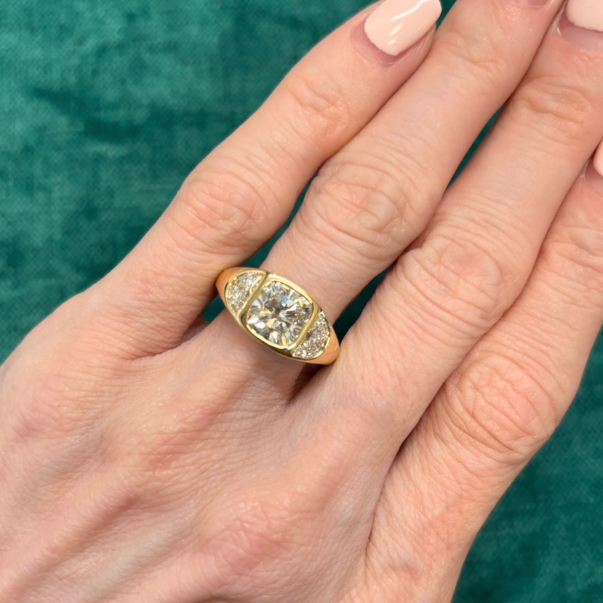 A hand with manicured nails wears the Burnish Set Cushion Diamond Engagement Ring with Half Moon Side Stones, showcasing a large cushion cut diamond in a bezel setting at the center, flanked by sparkling half moon side stones on each side, against a green background.
