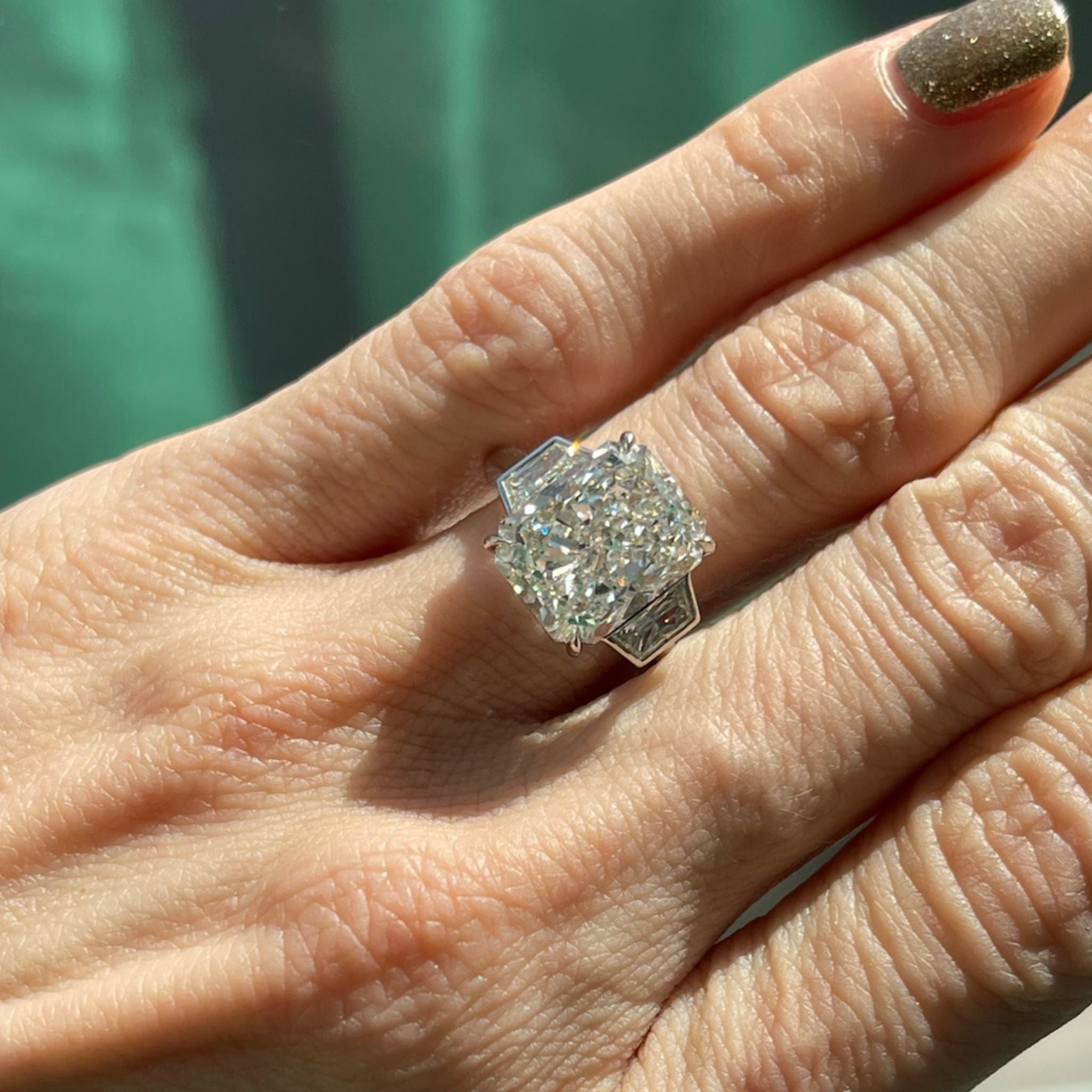 A close-up of a hand with a glittery gold fingernail wearing the Radiant Cut Diamond Engagement Ring with Bezel Set Epaulette Side Stones, set in silver, as sunlight highlights the ring’s sparkling brilliance.