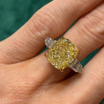 A hand wearing the Fancy Yellow Radiant Cut Diamond Engagement Ring with Tapered Baguette Side Stones, featuring a prominent yellow radiant-cut diamond at the center flanked by tapered baguette diamonds, set against a green background.
