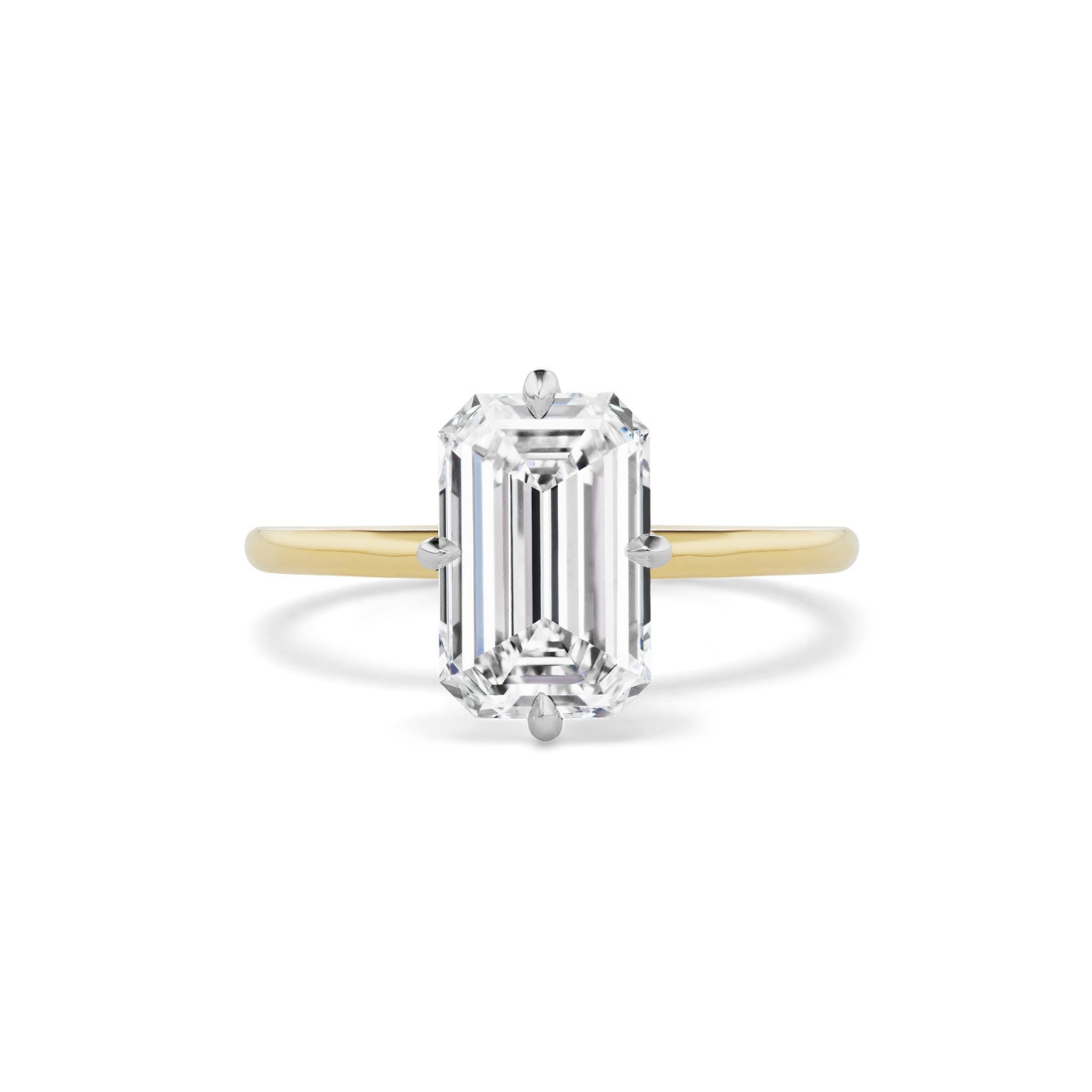 The Emerald Cut Diamond Engagement Ring with Compass Prongs features a large, emerald-cut clear diamond set in a simple prong setting on a gold band, elegantly photographed against a white background. This exquisite piece beautifully captures the essence of classic bridal rings.