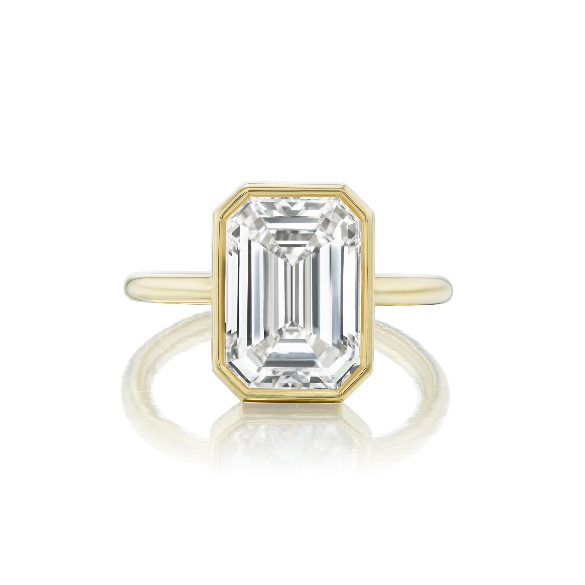 The Bezel Set Emerald Cut Diamond Engagement Ring showcases a stunning, emerald-cut diamond elegantly set in a minimalist 18k yellow gold bezel band, beautifully presented against a white background.