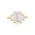 The Emerald Cut Diamond Engagement Ring with Trapezoid Side Stones showcases a central emerald-cut diamond, accented by two brilliant trapezoid side stones, all set in sophisticated 18k yellow gold prongs and elegantly displayed against a pristine white background.