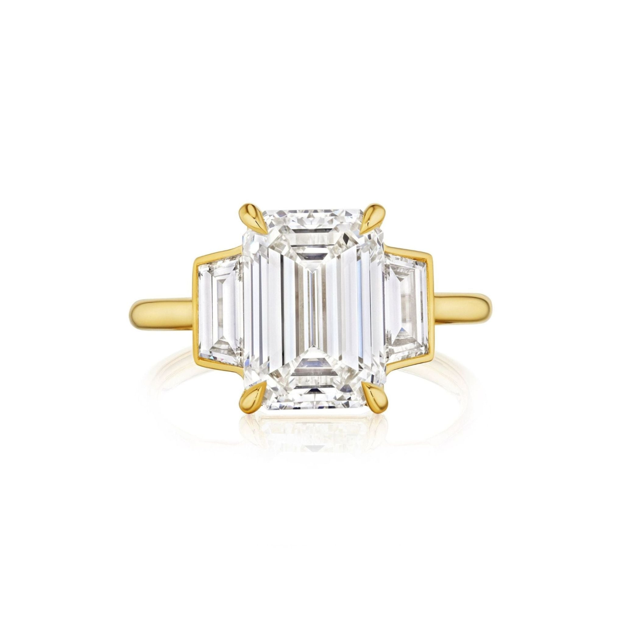 The Emerald Cut Diamond Engagement Ring with Trapezoid Side Stones showcases a central emerald-cut diamond, accented by two brilliant trapezoid side stones, all set in sophisticated 18k yellow gold prongs and elegantly displayed against a pristine white background.