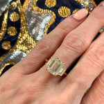 A hand with manicured nails wears the Emerald Cut Diamond Engagement Ring with Trapezoid Side Stones, showcasing a large emerald-cut diamond set in 18k yellow gold and flanked by elegant trapezoid diamond accents. The background displays blue fabric adorned with gold embroidery.