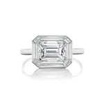 An East-West Set Mosaic Diamond Engagement Ring featuring an emerald-cut diamond with beveled corners, framed by a baguette halo and set on a simple band of white gold or platinum, displayed against a white background.