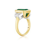 A Bezel Set Emerald Cut Emerald Engagement Ring in yellow gold and platinum, adorned with striking trapezoid baguette diamond side stones set in white metal, is displayed against a crisp white background.