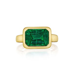 Introducing the East-West Bezel Set Emerald Cut Emerald Engagement Ring in Yellow Gold, featuring a vibrant emerald-cut emerald set horizontally at the center. The luxurious 18k yellow gold band gracefully enhances the gemstone's radiant green hue, creating a timeless and sophisticated look against a white background.