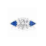 Introducing the Round Diamond Engagement Ring in White Gold with Kite Shape Blue Sapphire Side Stones – a breathtaking design featuring a brilliant round center diamond, accented by uniquely shaped kite blue sapphires on either side and set gracefully on a sleek white gold band.